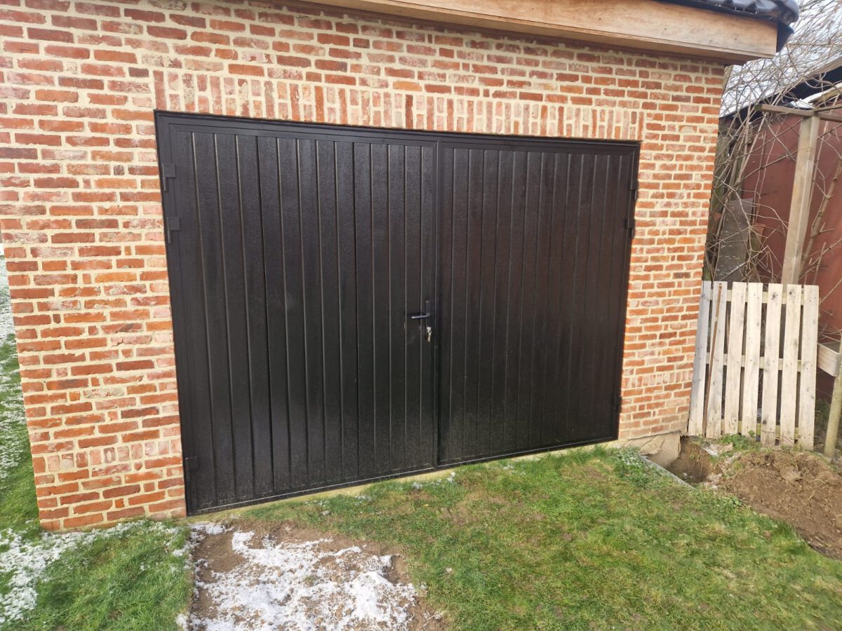 Een openslaande garagedeur met een full-line houtnerf structuur in RAL 9005 werd geïnstalleerd in een garage die zich achter in de tuin bevindt.