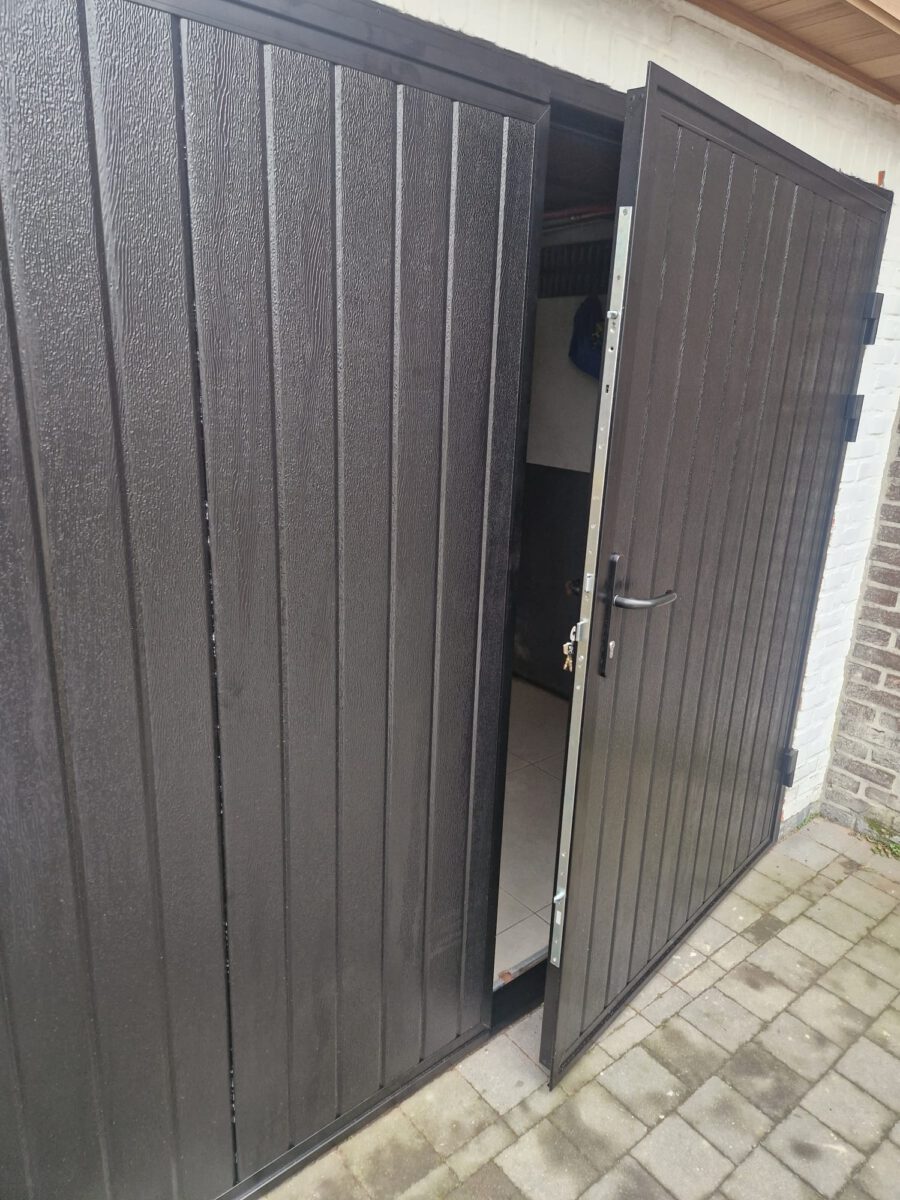 De openslaande garagedeur is uitgevoerd in een zwarte Full-Line houtnerf structuur met een verticale verdeling, conform RAL 9005. Daarnaast is een driepuntssluiting met bijbehorende sleutels zichtbaar.